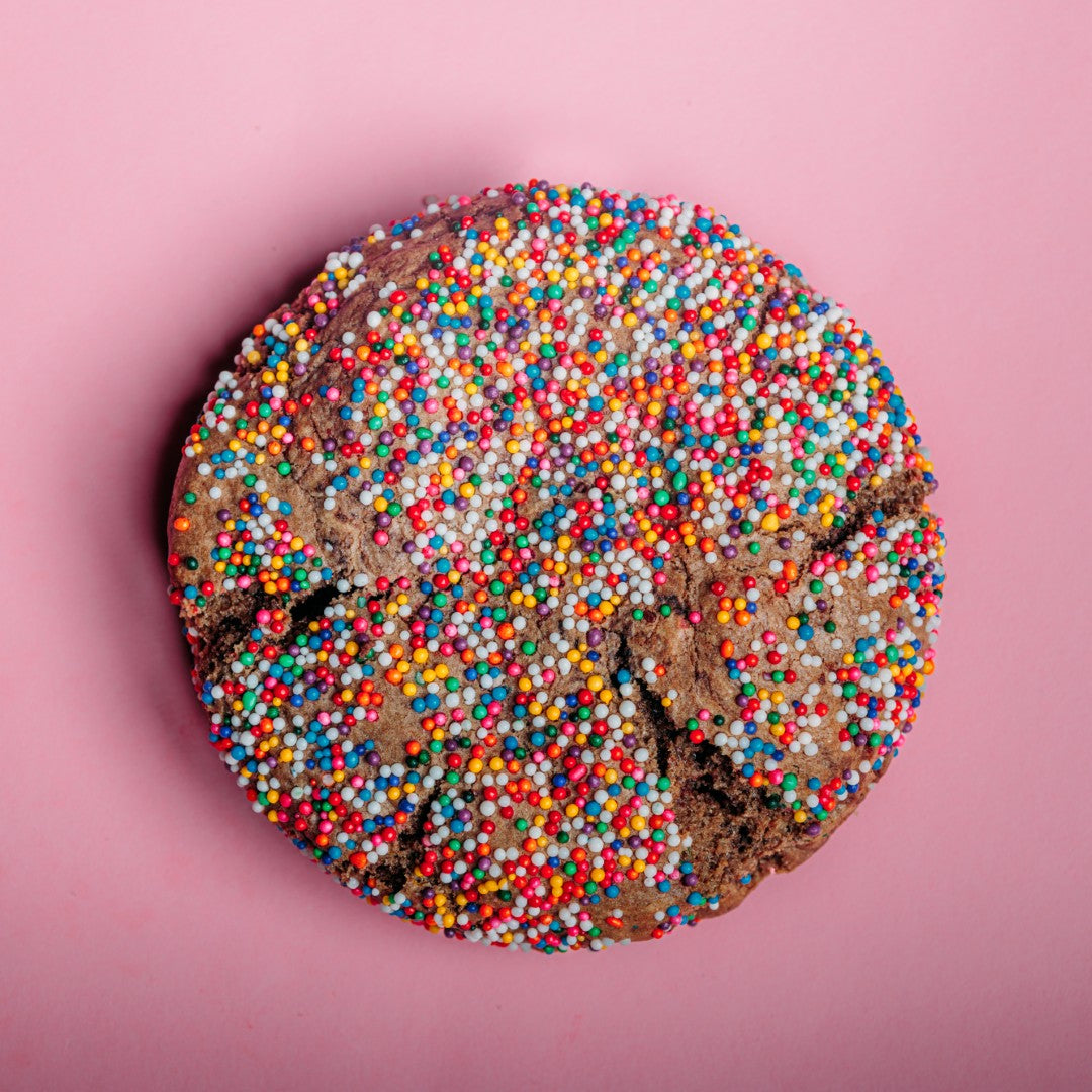 The Common Cookie's Chocolate Sprinkle Sugar Cookie - Gluten Free (Dozen) features a round cookie topped with colorful sprinkles, shown on a pink background.