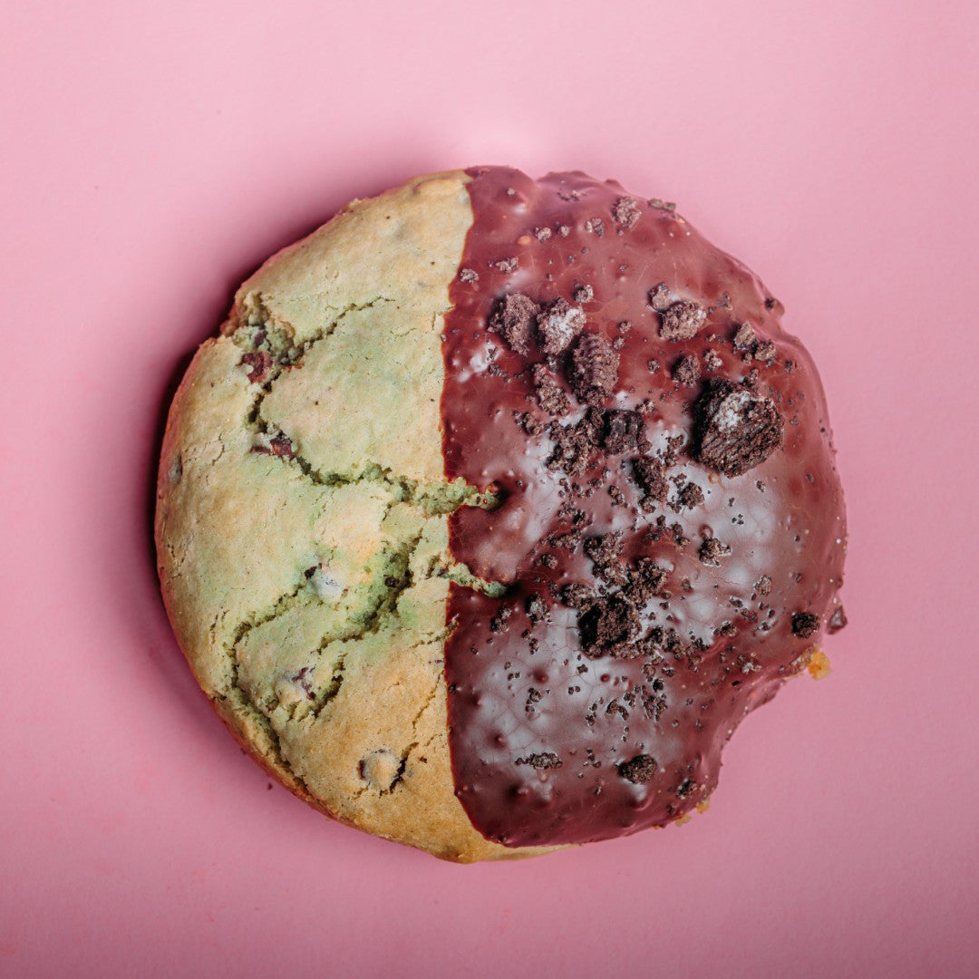 A large Grasshopper Pie Cookie - Gluten Free by The Common Cookie on a pink background, half plain and half coated in chocolate and cookie crumbles.