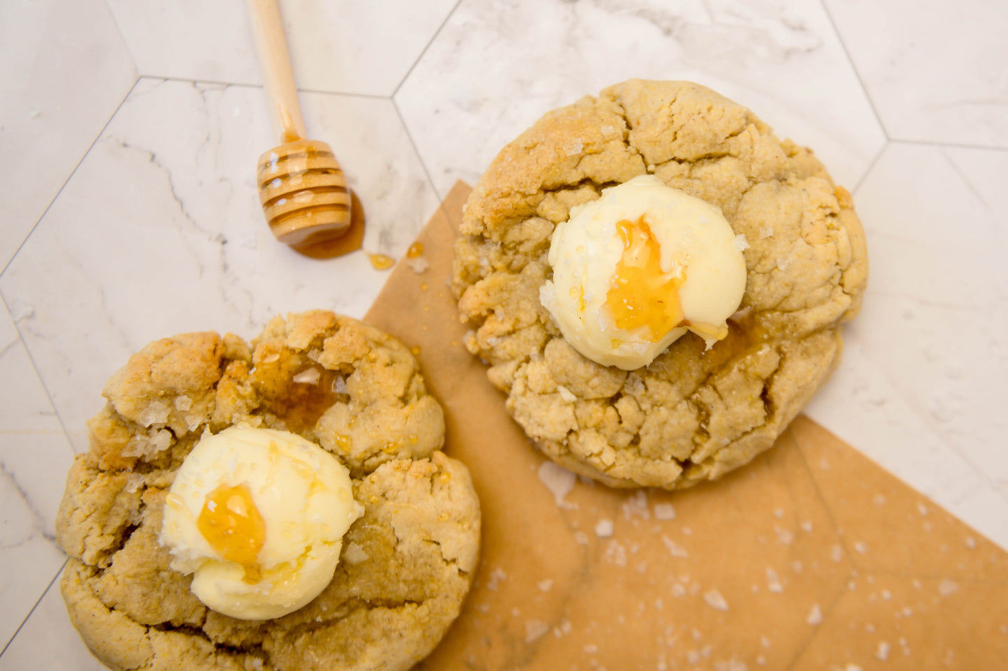 A dozen Salted Honey Cornbread - Gluten Free from The Common Cookie are topped with whipped honey butter and a drizzle of honey, served on parchment paper with a honey dipper on a white tiled surface.