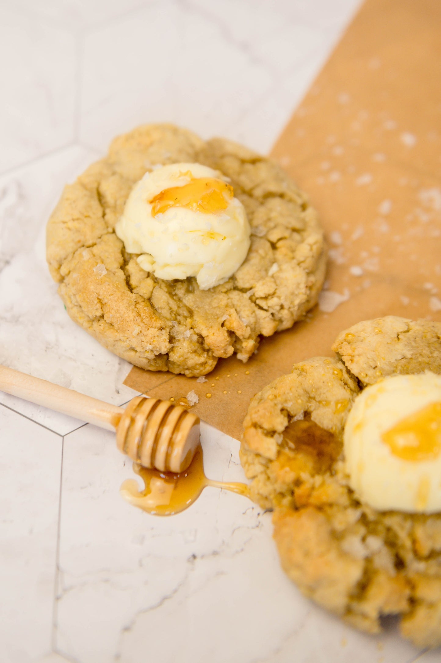 Two Salted Honey Cornbread - Gluten Free cookies by The Common Cookie, topped with whipped honey butter and a drizzle of honey, are served on parchment paper with a honey dipper beside them.