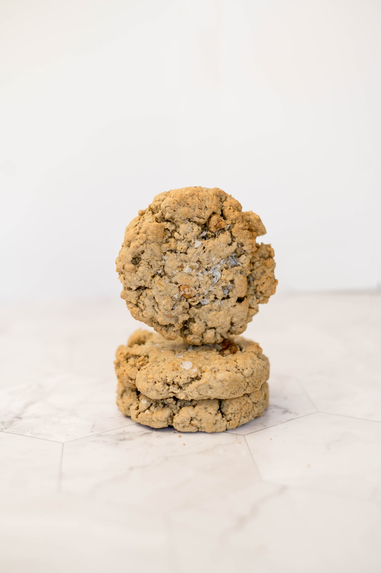 Two thick Dulce Salted Oatmeal - Gluten Free cookies from The Common Cookie, made with cinnamon oats, are stacked on a light marble-like surface against a neutral background.