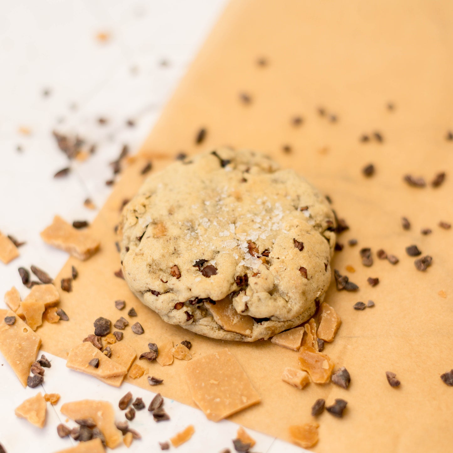 The Common Cookie's Cacao Nib with Coconut Toffee—Vegan and Gluten Free (Dozen)—rests on brown parchment, surrounded by chocolate chips and toffee pieces.