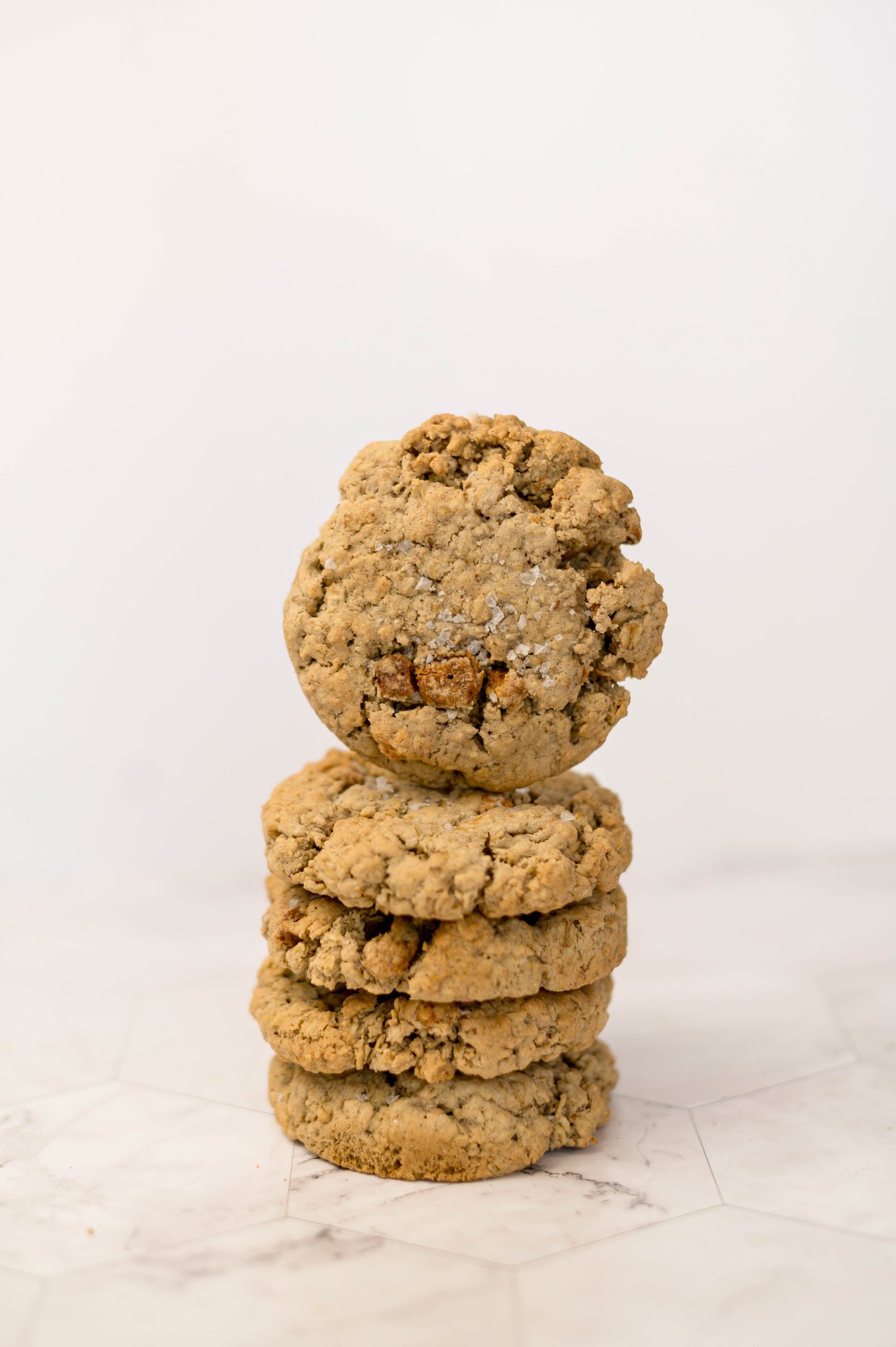 A stack of four The Common Cookie Dulce Salted Oatmeal - Gluten Free cookies, made with cinnamon oats, is shown against a white background, with the top cookie slightly bitten.