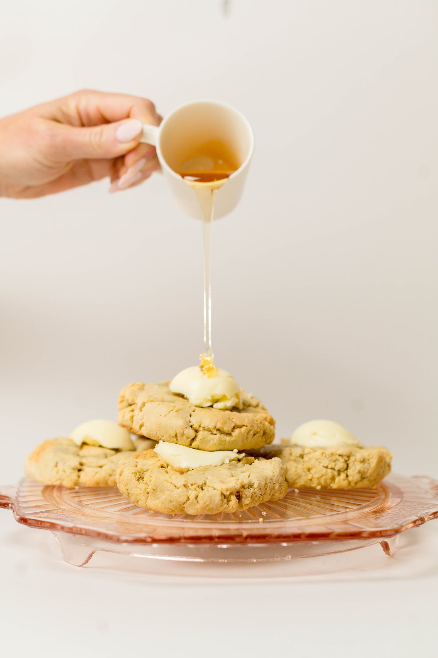 A hand pours honey onto The Common Cookie’s Salted Honey Cornbread - Gluten Free (Dozen), topped with whipped honey butter, served on a pink glass plate against a plain background.