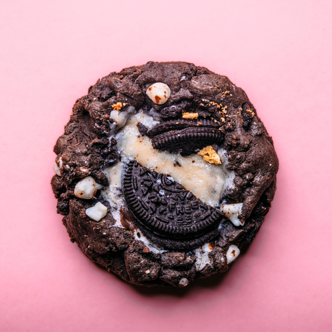 The Common Cookie’s Black Cocoa Oreo Cheesecake (Gluten Free, Dozen) features a chocolate cookie with white chocolate chips and a broken Black Cocoa Oreo on top, photographed on a pink background.