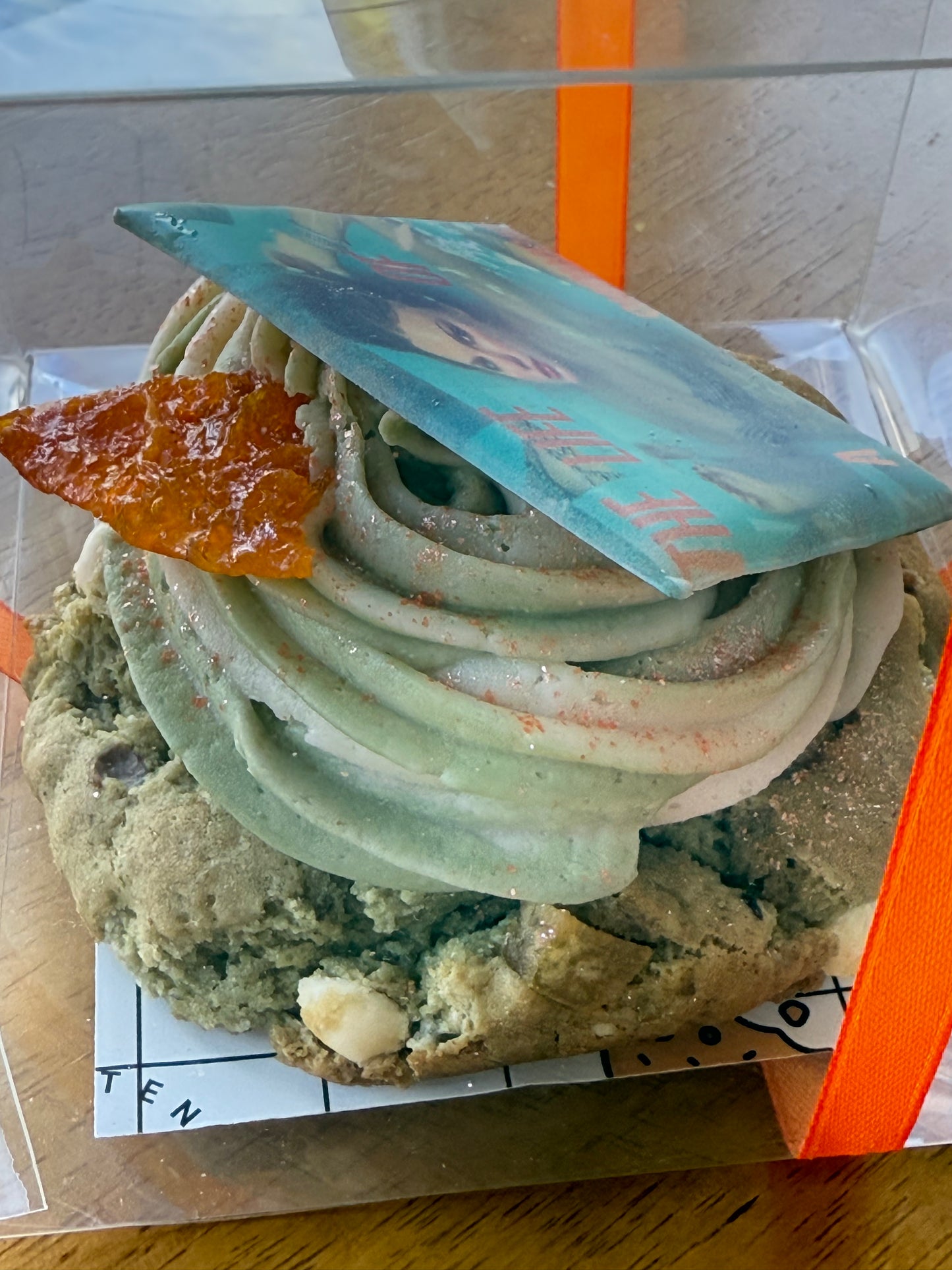 A Matcha Creamcicle-Gluten Free cookie by The Common Cookie, topped with green frosting, an edible printed image, and a small amber sugar shard, comes in a clear box tied with an orange ribbon.