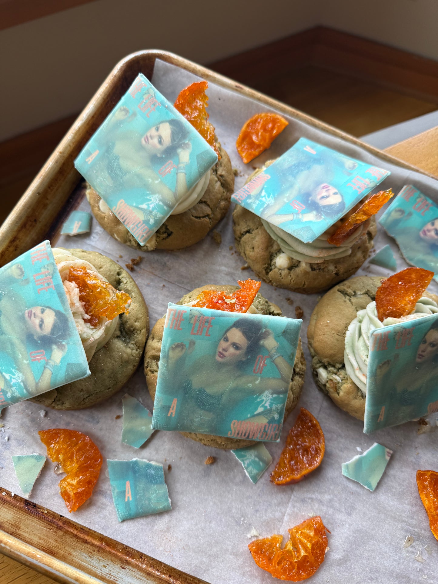 The Common Cookie's Matcha Creamcicle-Gluten Free features dulce salted oatmeal cookies with light green frosting, orange candied pieces, and edible printed squares with a woman's face on a parchment-lined tray. Gluten free.