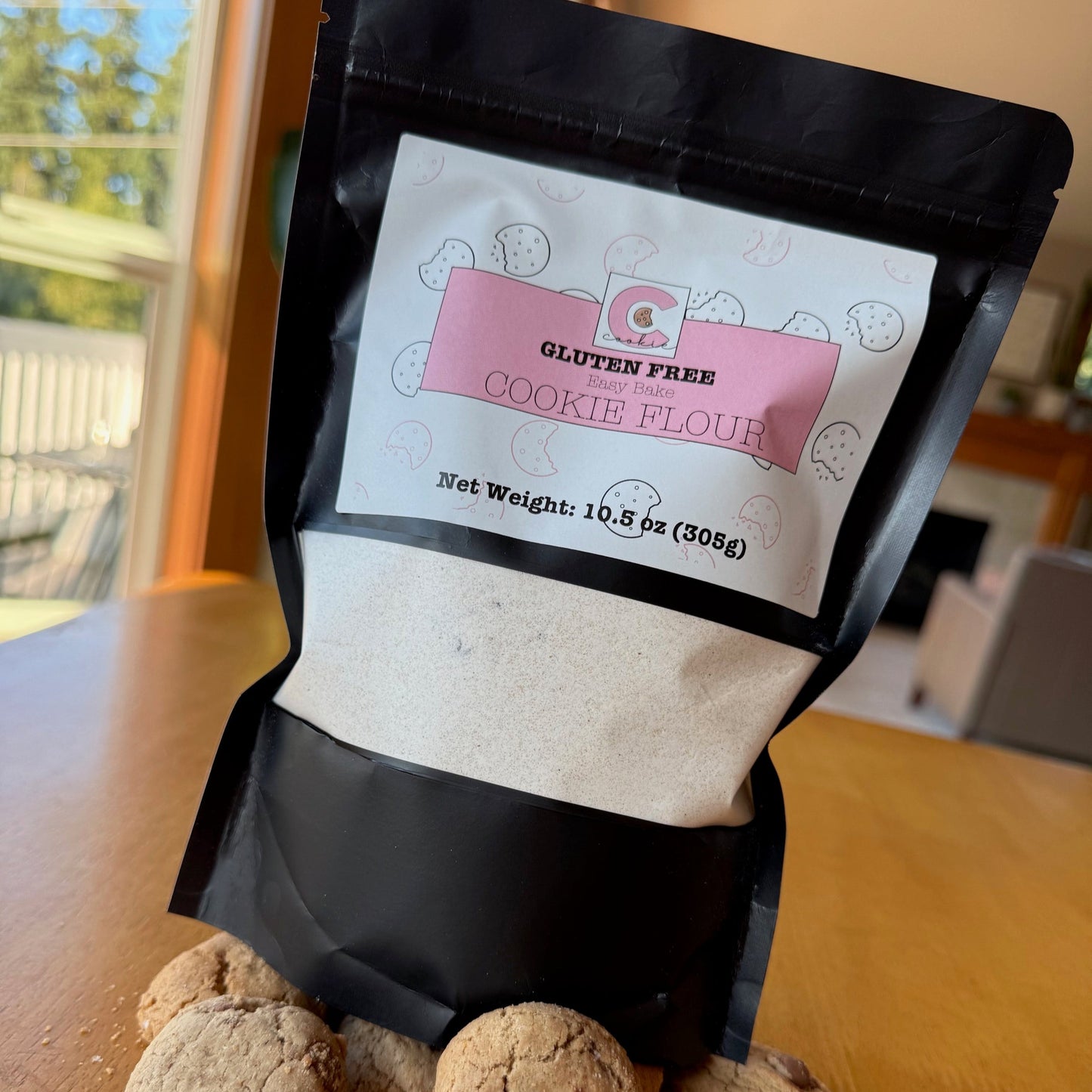 A bag of The Common Cookie's Bake Your Own Common Cookie mix sits on a wooden table, surrounded by chocolate chip cookies, one split in half.