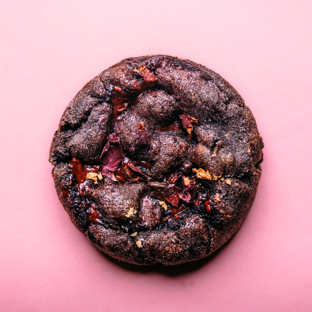The Common Cookie's Raspberry Rose Ganache Cookie-Gluten Free (Dozen) is a round, dark chocolate treat with Black Cocoa, topped with red and gold accents, displayed on a pink background.
