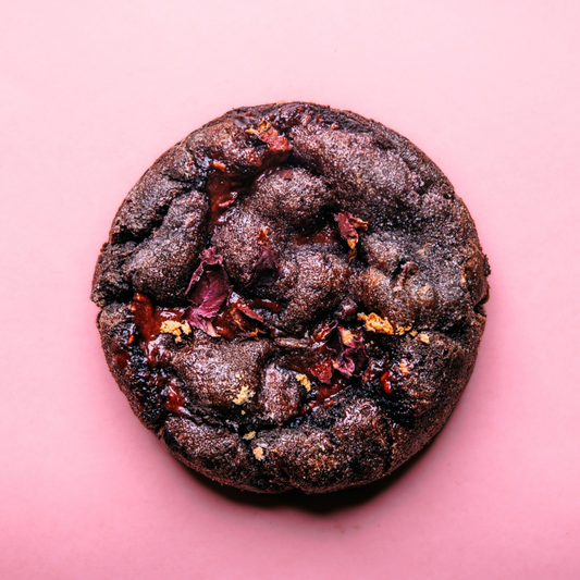 The Common Cookie's Raspberry Rose Ganache Cookie-Gluten Free (Dozen) is a round, dark chocolate treat with Black Cocoa, topped with red and gold accents, displayed on a pink background.