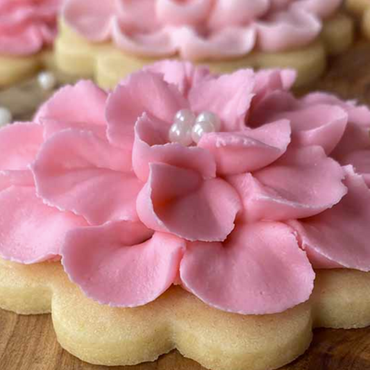 The Common Cookie's Mothers Day Flower Cookie—Gluten Free—features a sugar cookie topped with pink flower-shaped buttercream and white pearl-like decorations in the center, presented on a wooden surface.