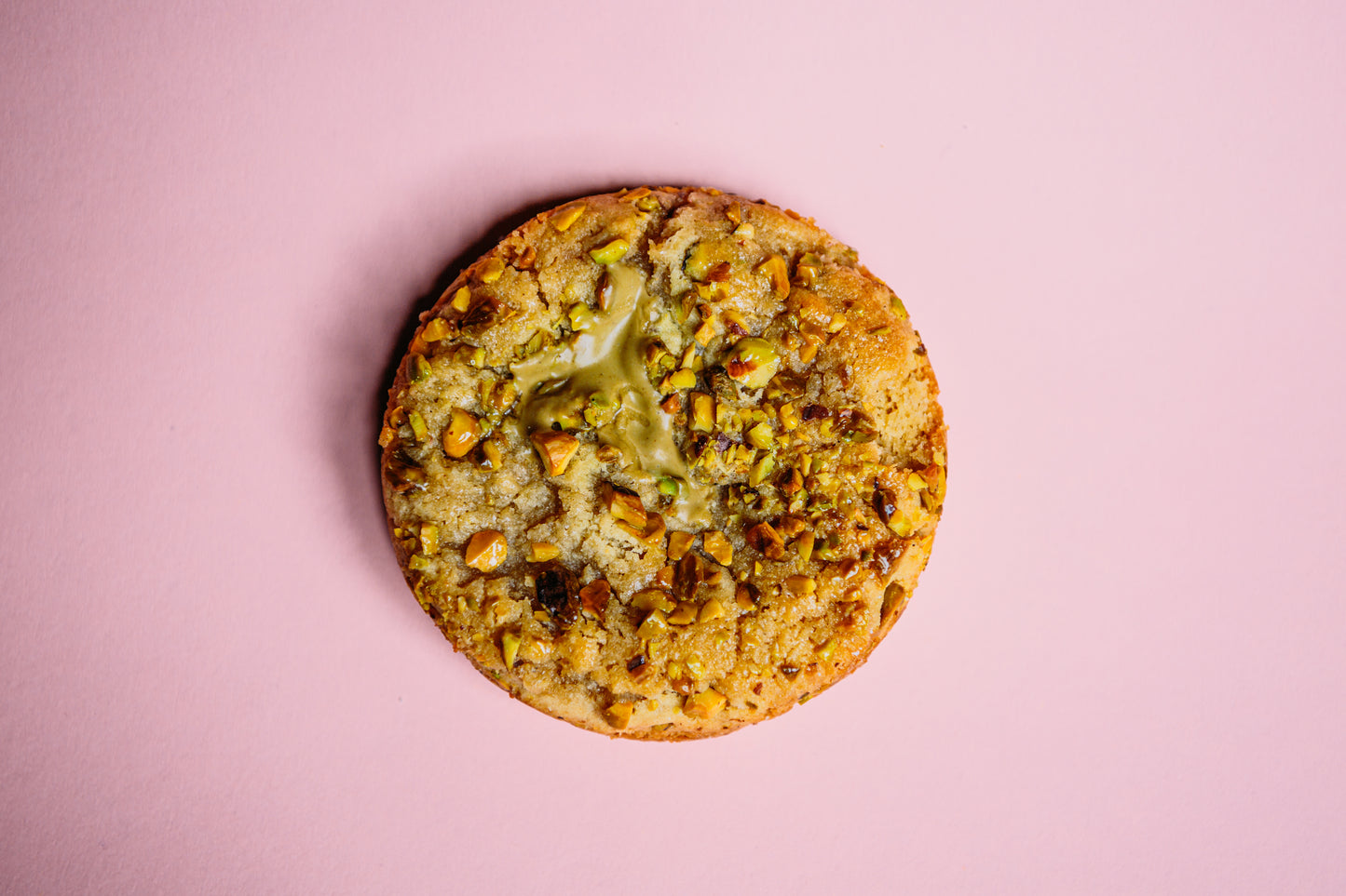Set against a pink background, The Common Cookie's Honey Pistachio Crunch - Gluten Free features crushed pistachios and luxurious pistachio cream filling.