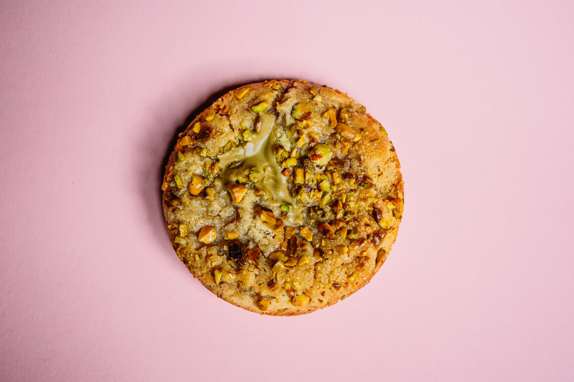 Set against a pink background, The Common Cookie's Honey Pistachio Crunch - Gluten Free features crushed pistachios and luxurious pistachio cream filling.