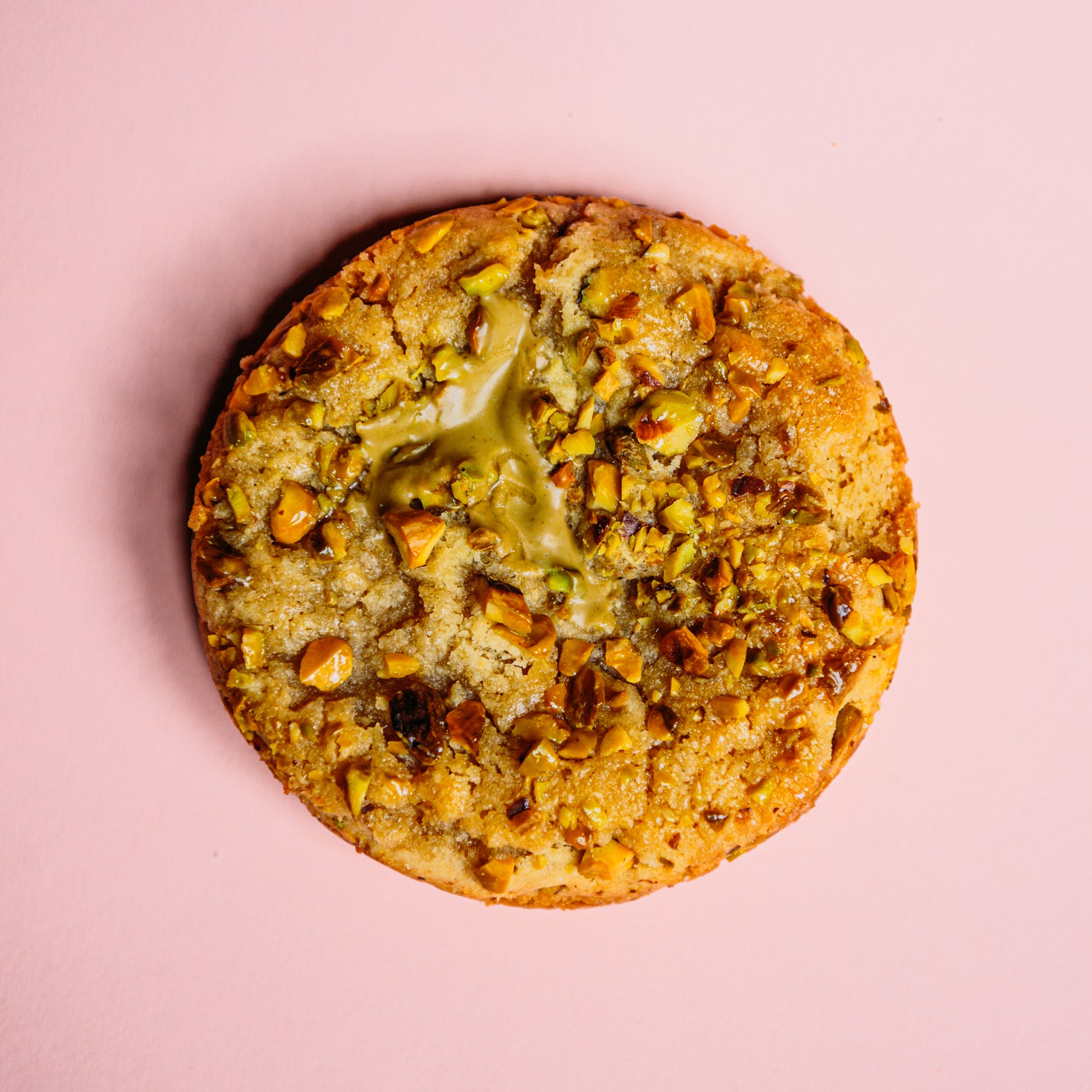 The Common Cookie's Almond Frangipane- Gluten Free pistachio cookie, with chopped nuts and a glossy glaze, rests on a pink background. Its buttery almond-scented dough makes it irresistibly delightful.