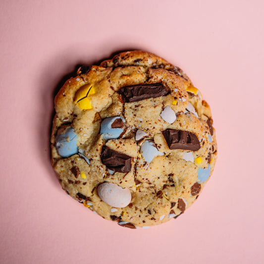 The Common Cookie's Almond Frangipane- Gluten Free features buttery almond-scented dough with chocolate chunks and colorful candy pieces, set against a pink background.
