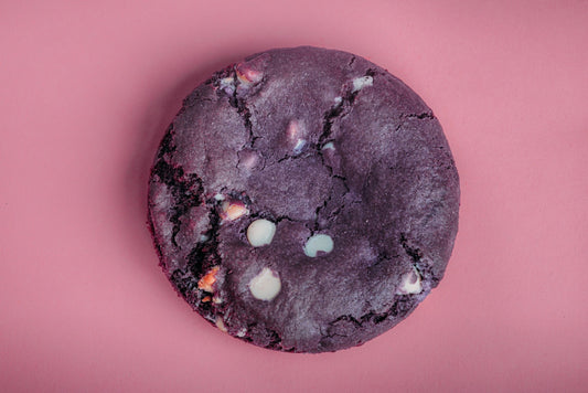 The Common Cookie’s Ube Stuffed Cheesecake Cookie- Gluten Free (Dozen), a round purple cookie with white chocolate chips, is displayed on a pink background.