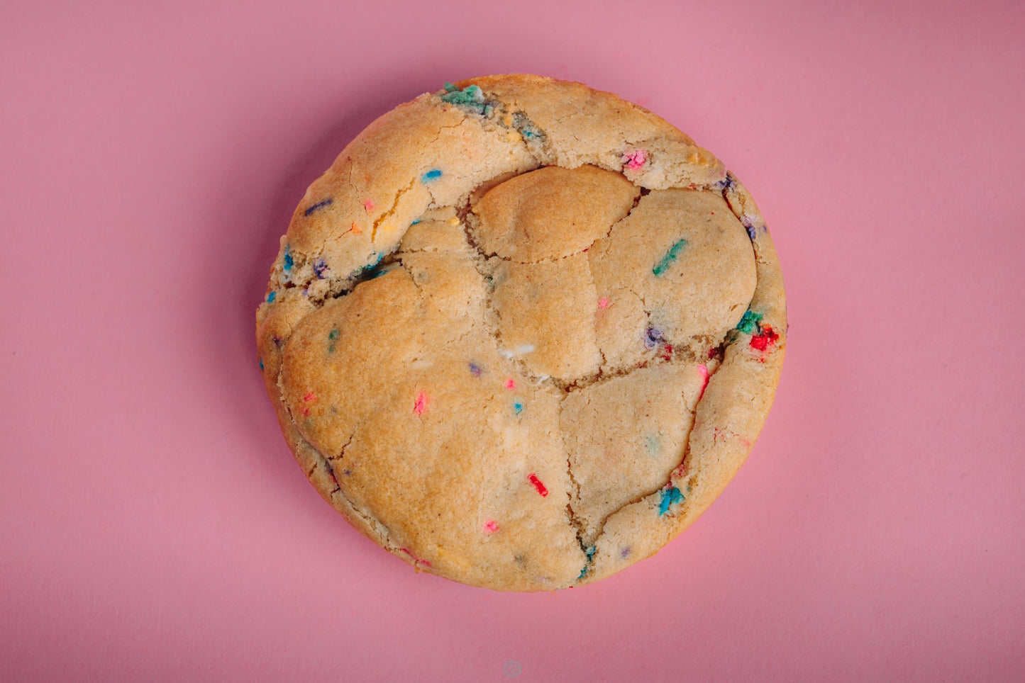 A large, round Birthday Cake Funfetti-Gluten Free cookie from The Common Cookie, topped with rainbow sprinkles, sits on a pink background and radiates colorful joy.