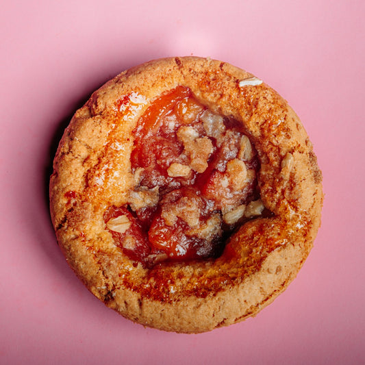 A gluten-free Peach Cobbler Cookie by The Common Cookie, featuring a crumbly crust and homemade flavor, topped with glazed peaches and photographed from above on a pink background.