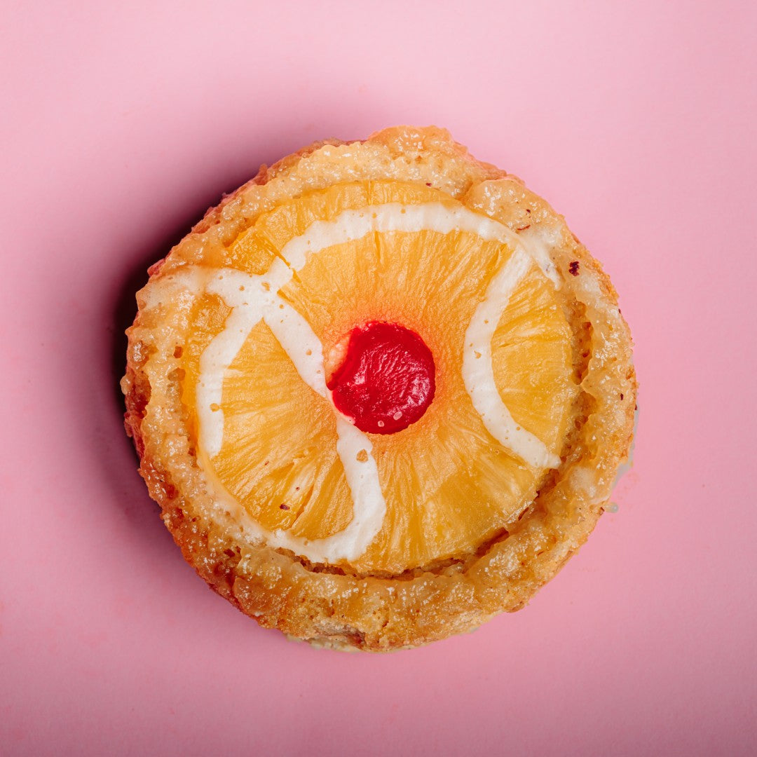 Enjoy a gluten-free Pineapple Upside-Down Cake Cookie by The Common Cookie, topped with pineapple, cherry, and vanilla glaze. Sold by the dozen—perfect for sweet cravings! Displayed on a pink background.