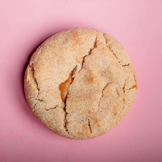 A round, cracked Dulce de Leche Snickerdoodle - Gluten Free cookie by The Common Cookie with a caramel center sits on a solid pink background.