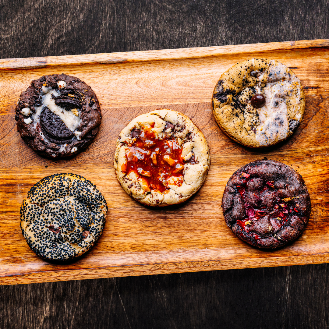 Display the *6 pack Bundle by Bundle—six assorted cookies with various toppings, arranged on a wooden tray against a dark backdrop—ideal for highlighting this tempting product on your Shopify store or as part of your Bundle Builder collection.