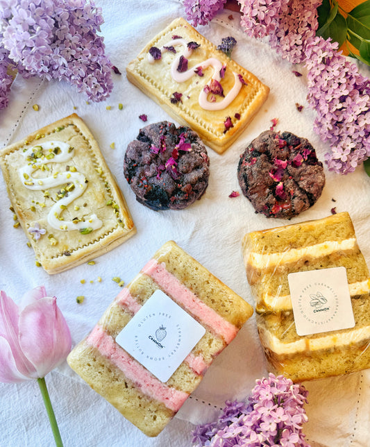 The Common Cookie’s Mothers Day Variety Pack features gluten-free pastries, cookies, and cakes arranged on a white cloth with lilac flowers and a pink tulip—a perfect handmade treat.