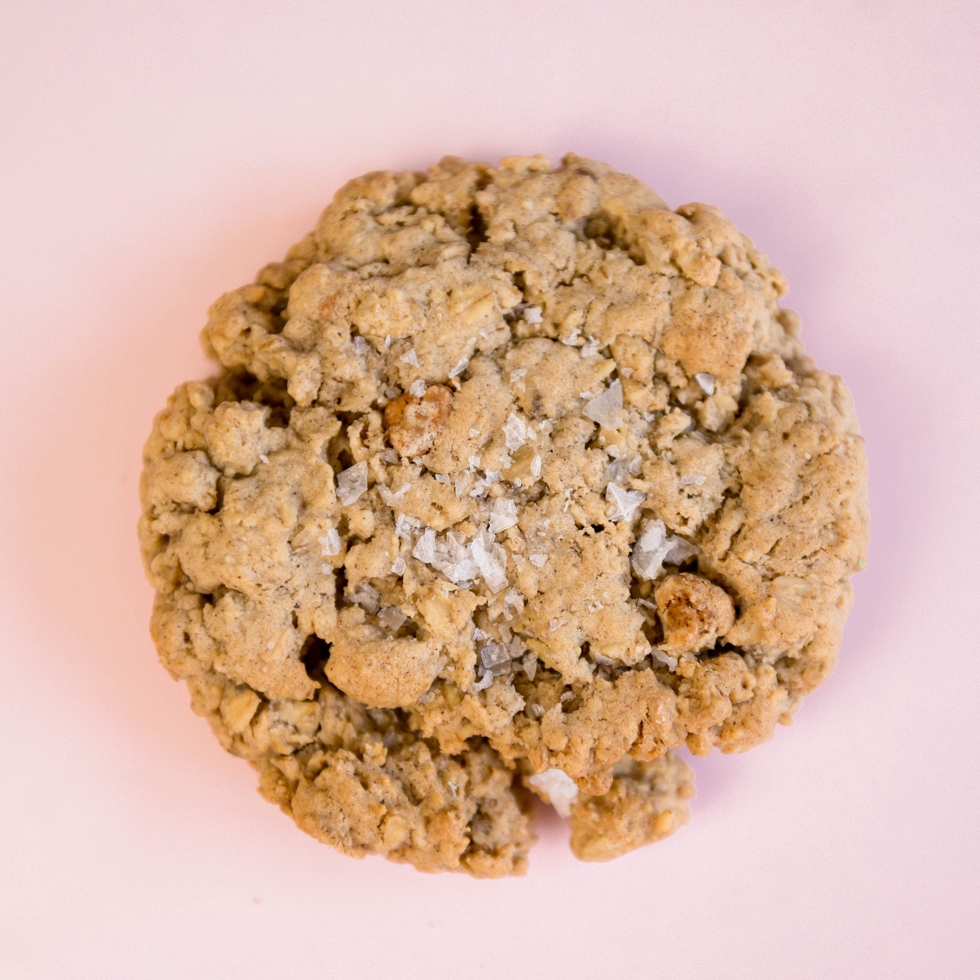 A single Dulce Salted Oatmeal - Gluten Free cookie from The Common Cookie, with a crumbly texture, sits on a light pink background.