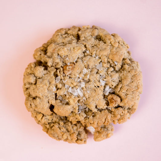 A single Dulce Salted Oatmeal - Gluten Free cookie from The Common Cookie, with a crumbly texture, sits on a light pink background.
