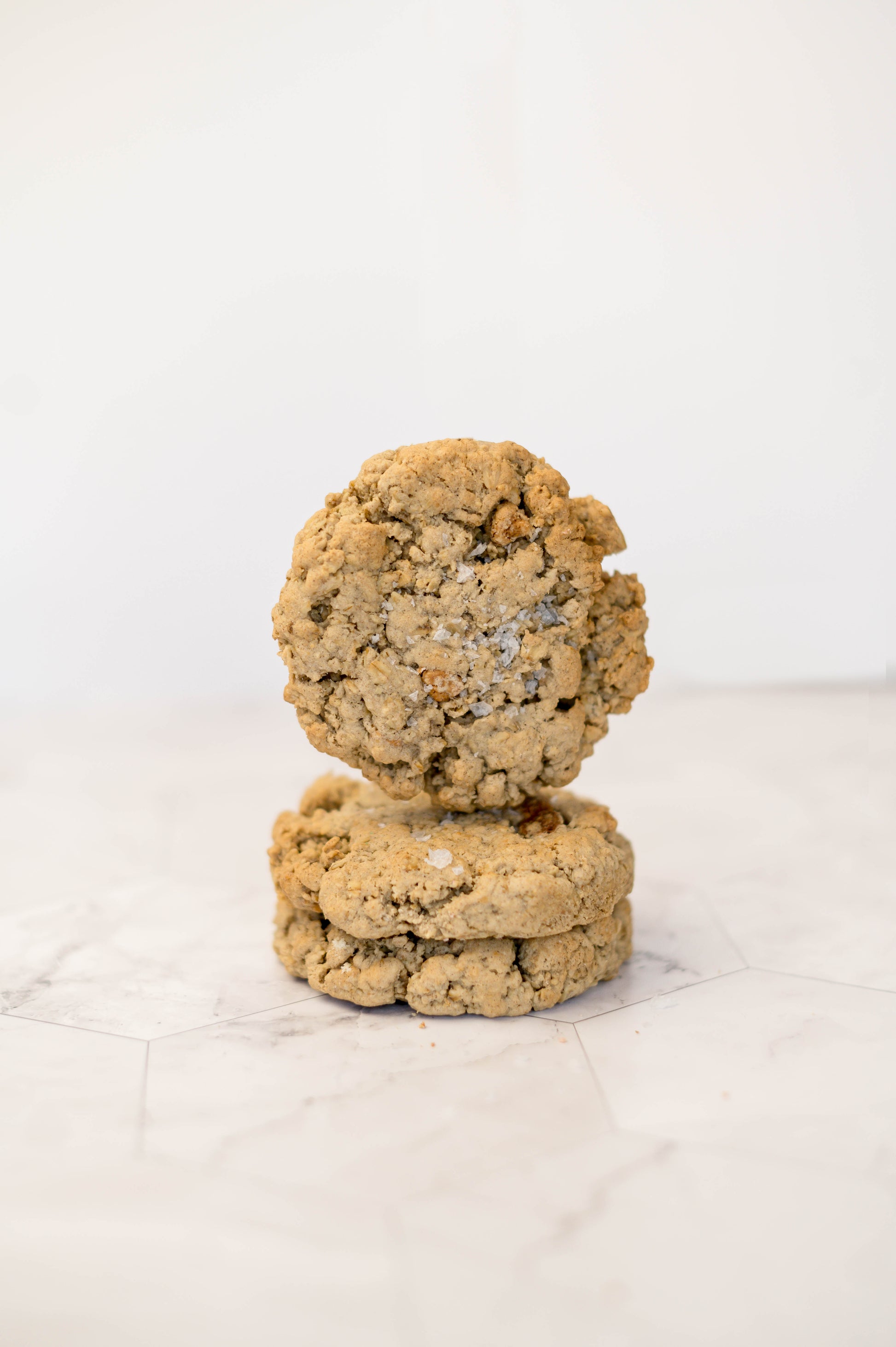 Two thick Dulce Salted Oatmeal - Gluten Free cookies from The Common Cookie, made with cinnamon oats, are stacked on a light marble-like surface against a neutral background.