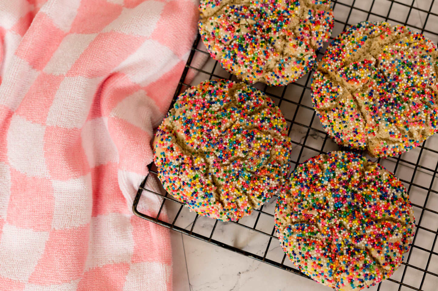 The Common Cookie's Dairy Free Sprinkledoodle - Gluten Free cookies, topped with rainbow nonpareils, cool on a rack next to a pink and white checkered cloth.