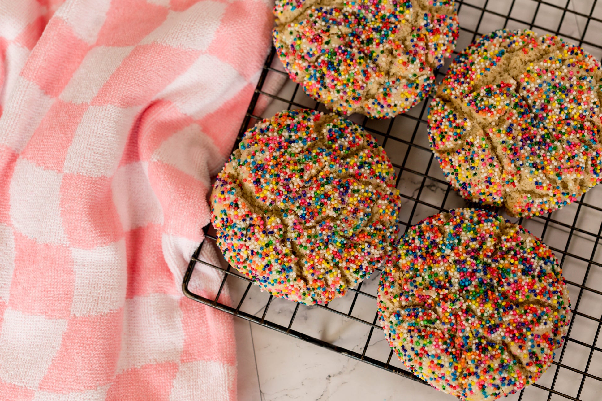 The Common Cookie's Dairy Free Sprinkledoodle - Gluten Free cookies, topped with rainbow nonpareils, cool on a rack next to a pink and white checkered cloth.
