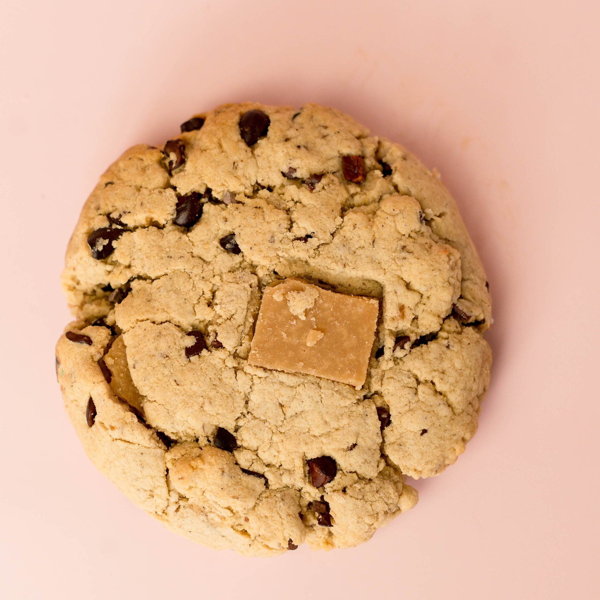 The Common Cookie’s Cacao Nib with Coconut Toffee—Vegan and Gluten Free (Dozen)—features a delicious, allergy-friendly cookie topped with light chocolate, shown on a pale pink background, perfect for crave-worthy snacking.