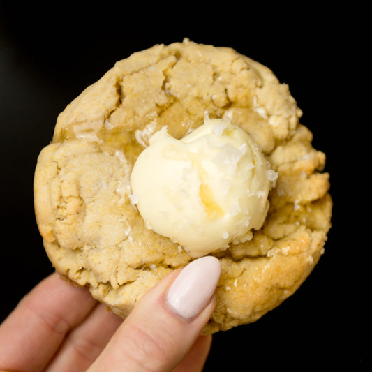 A hand holds The Common Cookie's Salted Honey Cornbread - Gluten Free cookie, topped with whipped honey butter, against a black background.