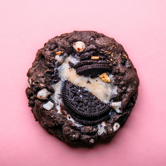 The Common Cookie’s Black Cocoa Oreo Cheesecake (Gluten Free, Dozen) features a chocolate cookie with white chocolate chips and a broken Black Cocoa Oreo on top, photographed on a pink background.