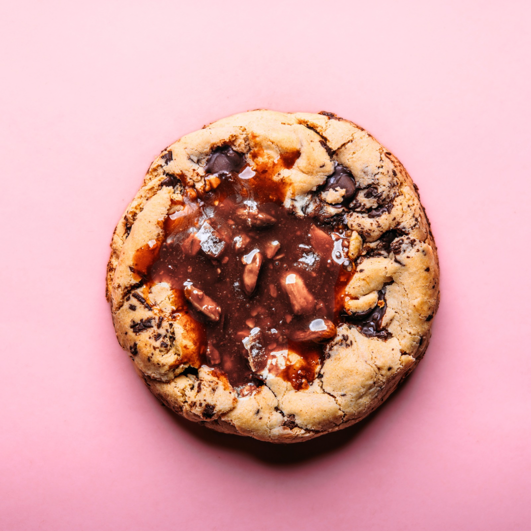 The Common Cookie's XL Stuffed Dubai Pistachio Quinoa Cookie— a gluten-free treat with chocolate chips and a pistachio-chocolate center— sits on a pink background.