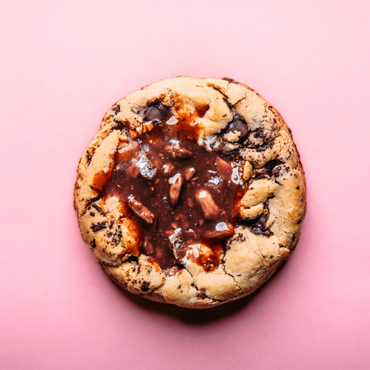 The Common Cookie's XL Stuffed Dubai Pistachio Quinoa Cookie— a gluten-free treat with chocolate chips and a pistachio-chocolate center— sits on a pink background.