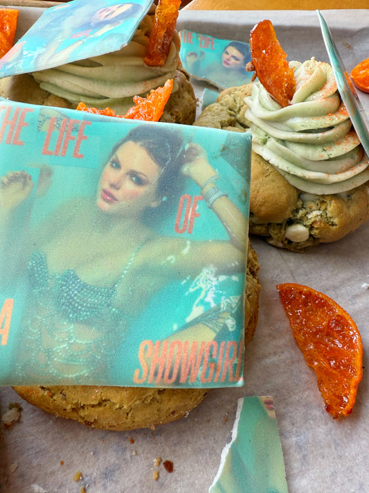 The Common Cookie’s Matcha Creamcicle-Gluten Free cookies topped with printed edible images of a woman underwater, green swirled frosting, white chocolate, and orange candied pieces arranged on parchment paper.