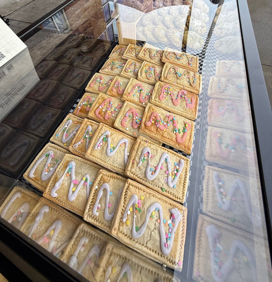 A dozen gluten-free Tar-Cookies by The Common Cookie, topped with icing and colorful sprinkles, are arranged in neat rows inside a glass bakery case.