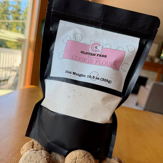 A bag of The Common Cookie's Bake Your Own Common Cookie mix sits on a wooden table, surrounded by chocolate chip cookies, one split in half.