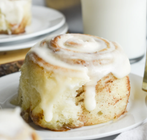 A close-up of The Common Cookie's Gluten Free Dozen Cinnamon Rolls, garnished with cream cheese frosting on a white plate and accompanied by a glass of milk in the background.