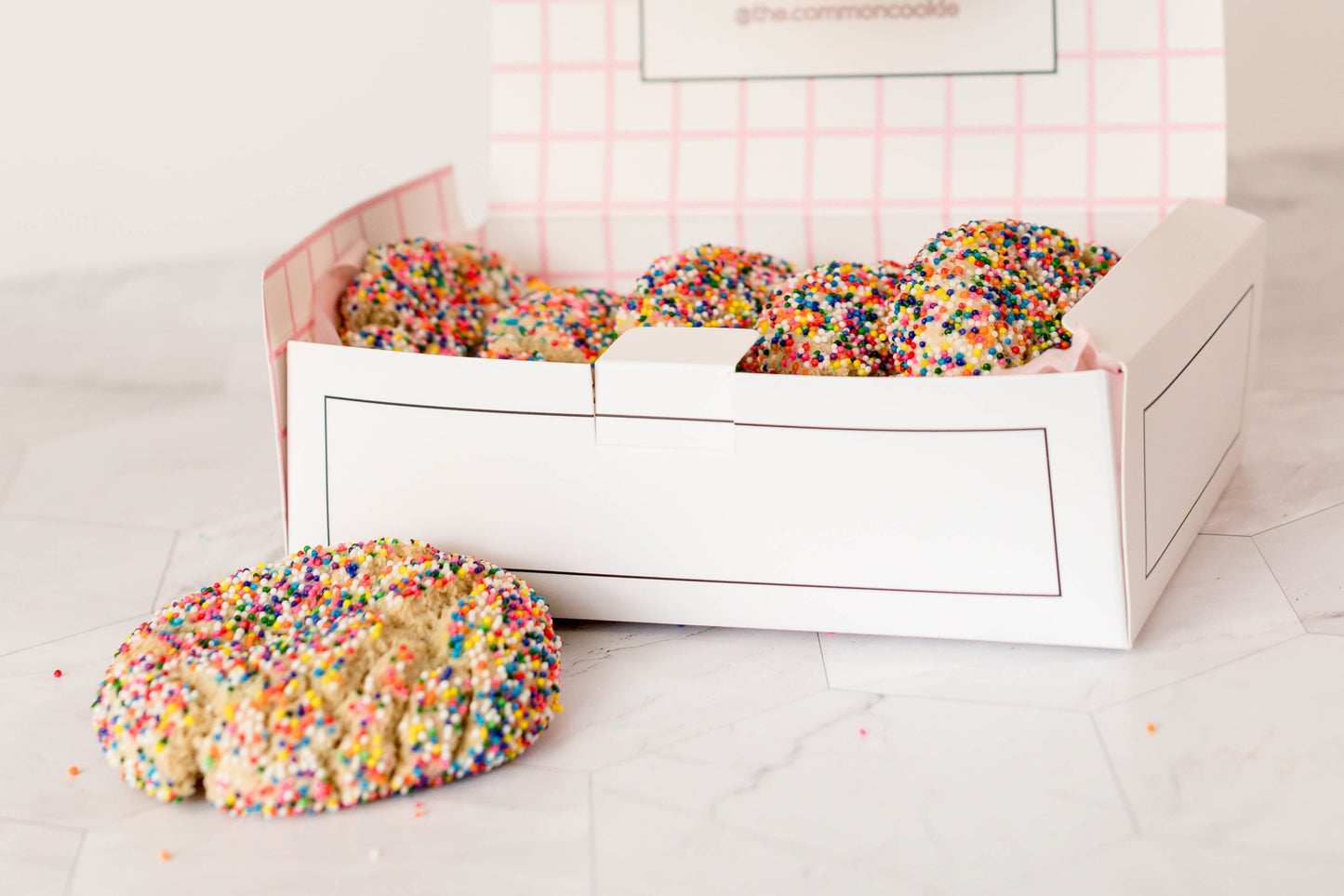 A 12-pack of The Common Cookie's treats, adorned with vibrant sprinkles, with one cookie displayed on the marble surface outside the box.